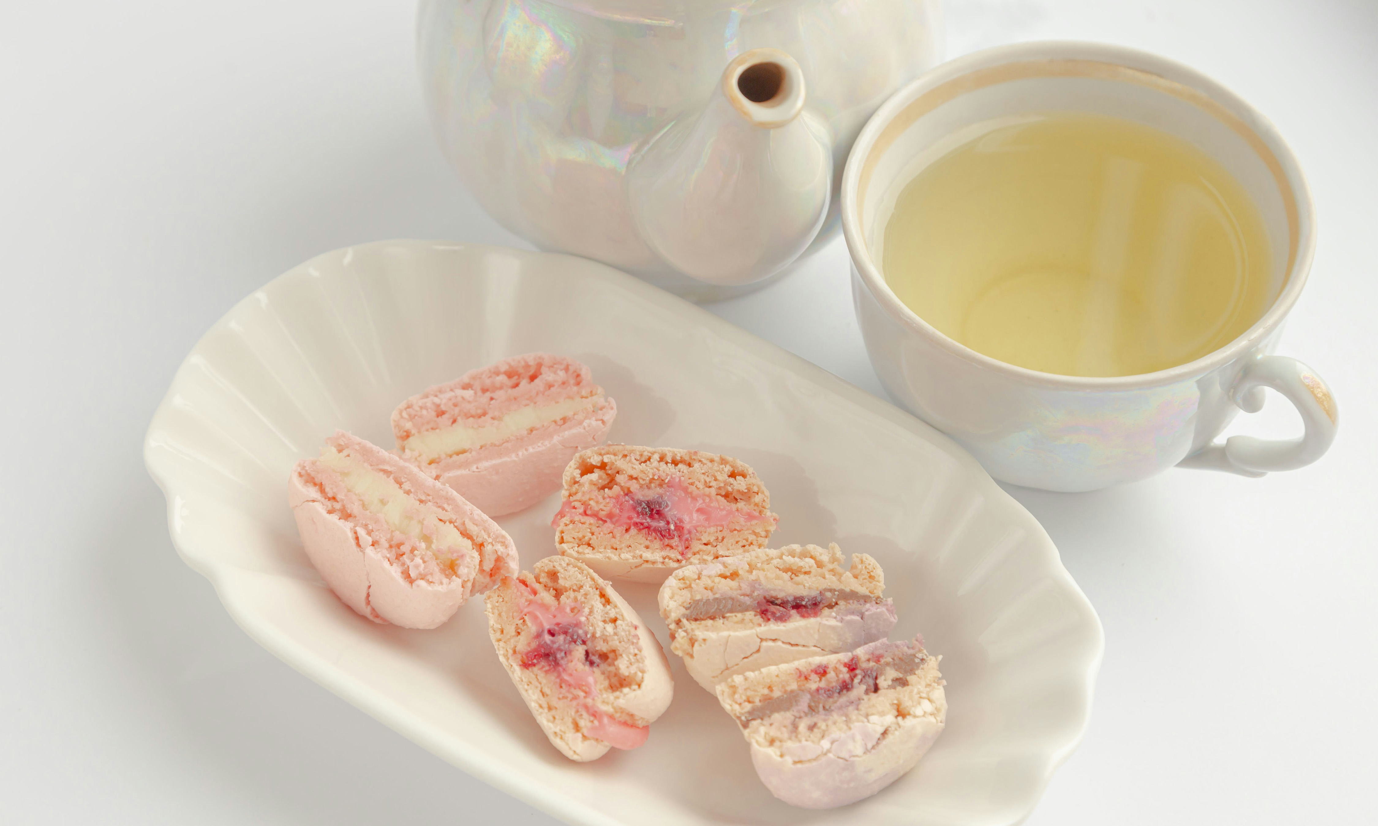 Three macarons cut in half on a plate next to a cup of tea and a teapot.