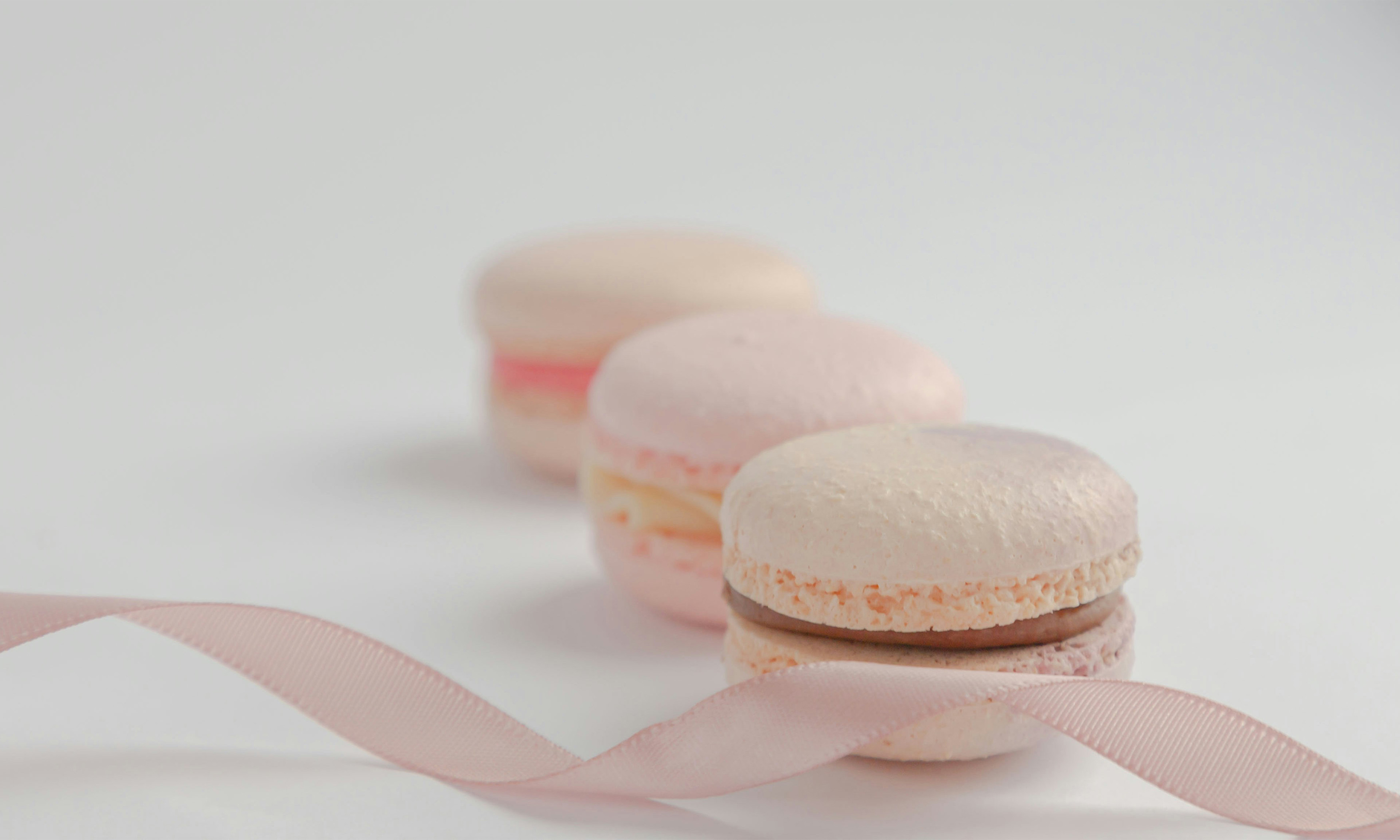 Three pink macarons arranged in a line moving away from the camera with a pink ribbon in front of the one closest to the camera.