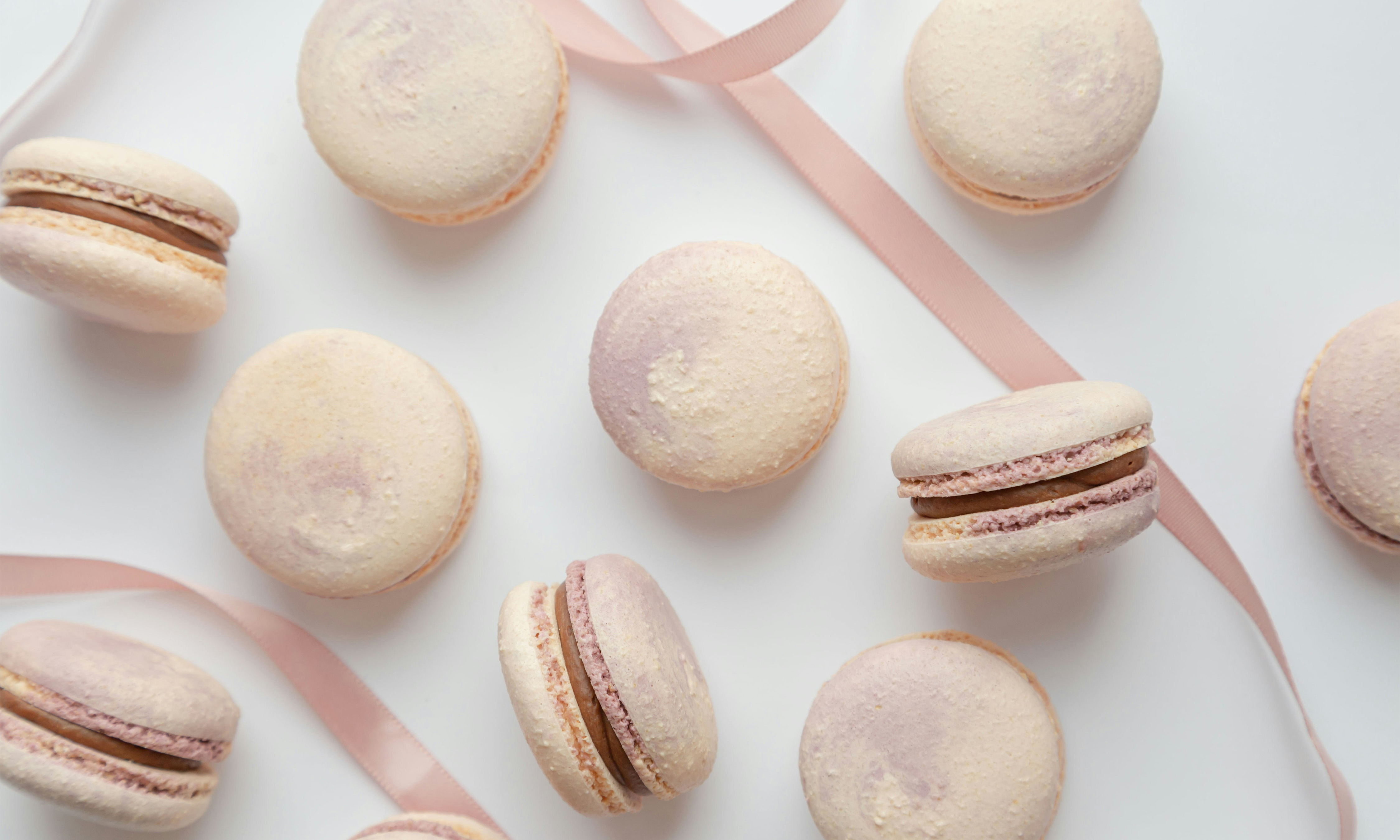Ten macarons, some flat and some standing up, scattered on a white surface with pink ribbon.