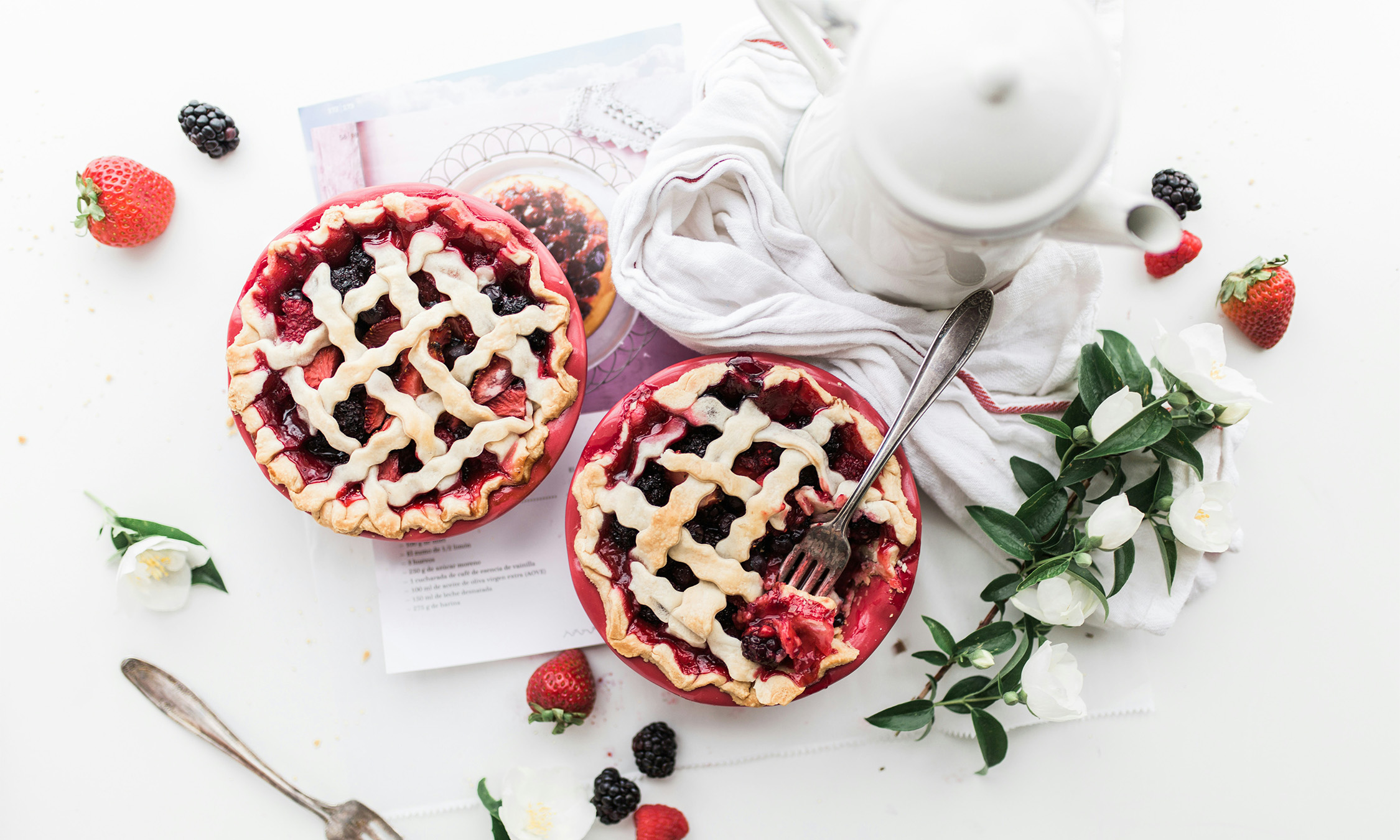Two berry pies on a table with blackberries and strawberries on the table surrounding them.